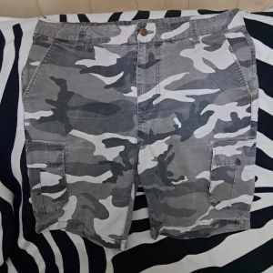 Grå camo cargoshorts med fickor - Snygga cargoshorts i grått camouflagemönster med flera praktiska fickor på sidorna och bak. Klassisk midja med bälteshällor och knappstängning. Använda endast en gång- väldigt bra skick:)