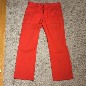 Röda chinos från Cross, W36 L32 - Snygga röda chinos från Cross i rak modell. Byxorna har klassisk passform med bälteshällor och fickor både fram och bak. Materialet är en mix av bomull, polyamid och elastan för lite stretch och skön känsla. Perfekta för dig som vill sticka ut med färg. Användes till golf. Liten grå fläck på högra benet där bak annars fläckfri!