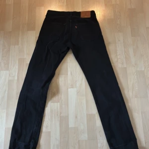 Svarta Levi's 501 jeans W31 L34 - Säljer ett par svarta Levi's 501 Premium jeans i klassisk rak modell. Jeansen har fem fickor, kopparfärgade nitar och är tillverkade i slitstark denim. Perfekta för dig som gillar tidlös stil och vill ha ett par riktigt snygga svarta jeans.