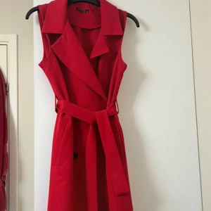 Röd ärmlös kappa med bälte - Snygg röd kappa med markerad krage, knappar framtill och matchande bälte i midjan. Ärmlös modell med stilren look och lätt utställd kjol. Perfekt för dig som vill sticka ut med en färgstark och trendig outfit. Dessutom så är den inte använd alls men det finns inget prislapp