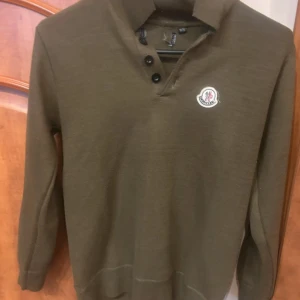 Olivgrön Moncler polotröja - Snygg olivgrön polotröja från Moncler med klassisk krage och tre knappar framtill. Tröjan har lång ärm och en broderad Moncler-logga på bröstet. Perfekt för dig som gillar stilrena och exklusiva plagg.