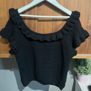 Svart stickad topp med volang - Supersöt svart stickad topp från H&M med bred volang runt halsringningen. Toppen har korta ärmar och en croppad passform som ger en trendig look. Perfekt att styla med höga jeans eller kjol för en fin look.💓