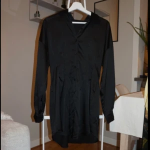 Svart kort skjortklänning i satin - Stilren svart skjortklänning med lång ärm och knappar framtill. Klänningen har en lätt glansig satinlook och markerad midja för en snygg siluett. Perfekt för dig som gillar minimalistisk och elegant stil.