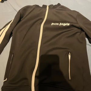 Palm Angels svart track jacket - Svart track jacket från Palm Angels med vita ränder längs ärmarna och vit dragkedja. Jackan har hög krage, två fickor med dragkedja och logga på bröstet. Perfekt för en sportig och trendig look.