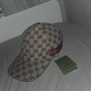 Gucci keps med GG-mönster och läderdetalj - Snygg keps från Gucci med klassiskt GG-monogram i beige och svart. Kepsen har justerbar rem i svart läder baktill och en ikonisk röd-blå rand på sidan. Tillverkad i bomullsmix med exklusiv känsla, perfekt för dig som vill sticka ut med lyxig streetstyle.
