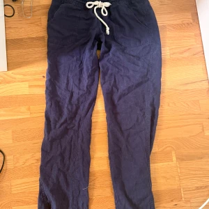 Mörkblå linnebyxor från Roxy - Snygga mörkblå linnebyxor från Roxy med avslappnad passform, elastisk midja och vit snörning. Byxorna har stora fickor fram och bak, perfekta för en chill och bekväm stil. Materialet är lätt och andas, perfekt för varmare dagar.