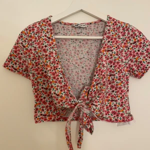 Blommig omlottopp  - Supersöt croppad omlottopp i storlek XS. Toppen har korta ärmar, v-ringning och knyt framtill. Mönstret är blommigt i rosa, rött, orange och grönt på vit botten. Perfekt för vår och sommar