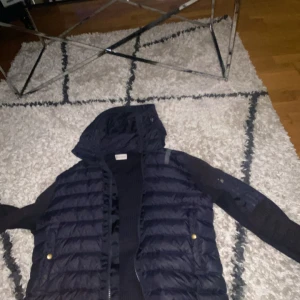 Mörkblå Moncler puffer med stickade ärmar - Snygg mörkblå pufferjacka från Moncler med stickade ärmar och huva. Jackan har dragkedja framtill, guldiga knappar på fickorna och en extra ficka med dragkedja på ärmen. Perfekt för dig som vill ha en unik och varm jacka med coola detaljer.all scann fungerar och jackan är äkta 