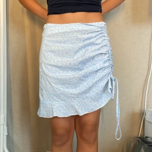 Blommig ljusblå kjol från SHEIN - Supersöt ljusblå kortkjol från SHEIN med vitt blommönster. Kjolen har snörning och rynk på ena sidan samt en liten volangkant nedtill. Perfekt för sommarens alla tillfällen och ger en fräsch vibe till din outfit.