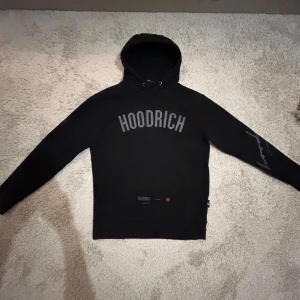 Svart hoodie från Hoodrich - Snygg svart hoodie från Hoodrich med tryck på bröstet och ärmen. Den har en klassisk passform med långa ärmar och en bekväm huva. Skicka 8/10, storlek XS men passar alla som har S, lite sliten vid höger armbåge.  Priset kan diskuteras! 