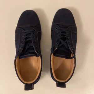 Mörkblå mockasneakers med röd sula (loubs) - Snygga sneakers i mörkblå mocka med svarta snören och cool röd sula. Skorna har rund tå, platt sula och diskreta detaljer. Insidan är ljusbeige och de har en klassisk, stilren look som passar till det mesta.