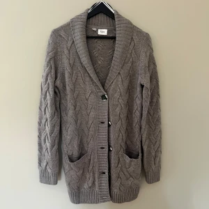 Grå stickad kofta från bpc - Mysig grå kofta från bpc med grovt flätstickat mönster, djup sjalkrage och stora knappar framtill. Koftan har två praktiska fickor och långa ärmar. Perfekt för lager-på-lager under kyliga dagar.