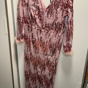 Rosa paljettklänning med fjädrar - Glittrig rosa långklänning med paljetter i olika rosa och röda nyanser. Djup v-ringning och långa ärmar med fluffiga fjädrar vid ärmsluten. Perfekt för dig som vill sticka ut och glänsa på festen.