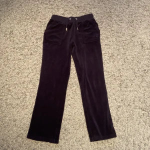 Mörk blå mjukisbyxor i sammet Juicy Couture - Mörk blå mjukisbyxor från Juicy Couture i mjuk sammet. Byxorna har raka ben, snörning i midjan, stora fickor fram och bak med knappar samt broderad JC-logga framtill. Perfekta för chill dagar och snygg streetstil.