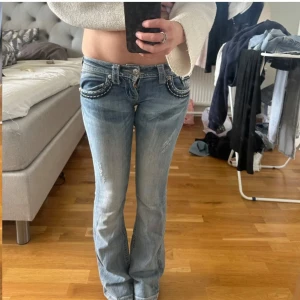 Miss Me ljusblå bootcut jeans med detaljer - Säljer ett par ljusblå jeans från Miss Me med låg midja och bootcut. Jeansen har slitna detaljer, broderade bakfickor med nitar, paljetter och dekorativa knappar. Snygga kontrastsömmar och coola detaljer som verkligen sticker ut. Perfekta för dig som gillar statement-jeans. Öppningar vid skrevet, lagningar längs benen fast syns knappast. Sytt mer lågmidjade