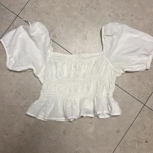 Vit blus med puffärm från Gina Tricot - Supersöt vit blus från Gina Tricot med puffiga korta ärmar och smockad överdel. Blusen har volangkant nedtill