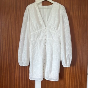 Vit broderad kortklänning med knappar - Superfin vit kortklänning med broderade blommor, v-ringning och långa puffärmar. Klänningen har knappar framtill och dragkedja bak. Perfekt för sommarens alla tillfällen och ger en romantisk vibe. Jättefin, endast testad 