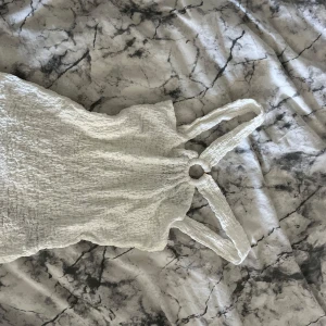 Vit smockad linnetopp med ringdetalj - Supersöt vit linnetopp med smockat tyg och breda axelband som möts i en cool ringdetalj framtill. Perfekt för varma dagar och enkel att styla med jeans eller kjol. Toppens struktur ger en trendig och chill vibe.