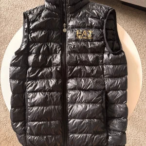 EA7 Emporio Armani väst  guldlogga som ny äkta - Säljer min EA7 Emporio Armani väst med guldfärgad logga. Äkta och i toppskick använd tre gånger. Inga repor eller skador. Originalpåse och EA7 kedja följer med. Storlek S. 