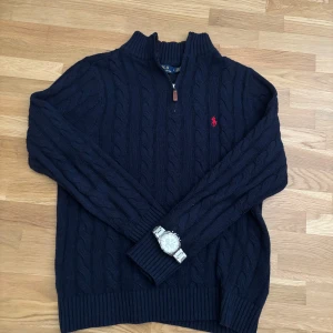 Mörkblå stickad tröja Polo Ralph Lauren - Säljer en mörkblå stickad tröja från Polo Ralph Lauren med kabelstickat mönster och röd logga på bröstet. Tröjan har hög krage och half zip-dragkedja, samt långa ärmar och ribbade muddar. Perfekt för dig som gillar klassisk och stilren look.