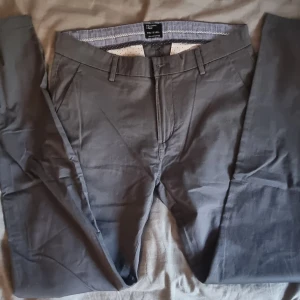 Grå slim chinos i ekologisk bomull - Snygga grå chinos i slim fit med klassisk design och bälteshällor. Tillverkade i ekologisk bomull för en skön känsla och stilren look. Perfekta för dig som gillar en smalare passform och vill ha ett par stilrena byxor till vardags.
