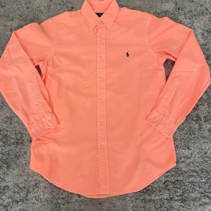 Korallfärgad skjorta från Ralph Lauren - Snygg korallfärgad skjorta från Ralph Lauren med klassisk krage och knäppning framtill. Skjortan har lång ärm och ikonisk blå broderad logga på bröstet. Tillverkad i mjuk bomull och passar perfekt för en fräsch och stilren look. Storlek S, hör av dig vid fundering