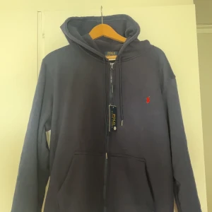 Mörkblå hoodie från Polo Ralph Lauren - Snygg mörkblå hoodie från Polo Ralph Lauren med dragkedja och huva. Klassisk röd broderad logga på bröstet och känguruficka framtill. Perfekt för en chill och stilren look. Tillverkad i mjuk bomull för extra komfort.
