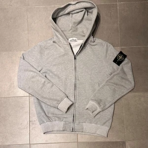 Grå hoodie från Stone Island - Säljer en stilren grå hoodie från Stone Island med klassisk logga på ärmen som är i bra skick. Tröjan har dragkedja framtill, huva och ribbade muddar. . Materialet är mjuk bomull. Passform till runt 170cm