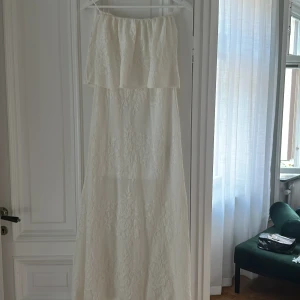  Vit volang långklänning i spets - Superfin vit långklänning med axelbandslös design och vacker spetsdetalj. Klänningen har en volang upptill. Perfekt skick (prislappen sitter kvar). 