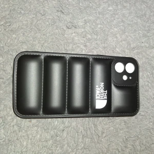 Svart The North Face mobilskal - Säljer en svart mobilskal från The North Face med vadderad design och logga på framsidan. Den har en rektangulär form och är tillverkad i syntetmaterial. Perfekt för att skydda mobilen och ge en cool streetstyle-vibe.