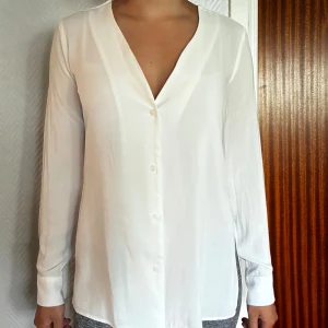 Ny vit v-ringad skjorta från Ivyrevel - Säljer en stilren vit skjorta från Ivyrevel med v-ringning och knappar framtill. Skjortan har lång ärm, slits i sidorna och är tillverkad i ett mjukt, lätt material som ger ett snyggt fall. Ej använt.