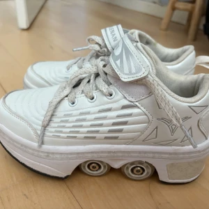 Vita sneakers med inbyggda hjul - Unika vita sneakers från Ainman med inbyggda hjul i sulan, perfekta för att rulla fram med stil. Skorna har snörning, kardborreband och coola silverdetaljer på sidorna. Ovandelen är i syntetmaterial och designen är sportig och futuristisk.