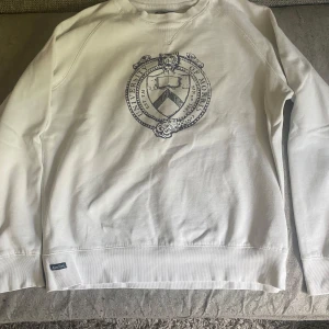 Vit sweatshirt från Morris Gents - Vit sweatshirt från Morris Gents med stort tryck av ett universitetsinspirerat emblem på bröstet. Tröjan har rund hals, långa ärmar och ribbade muddar. Perfekt för en chill och stilren look. Passar M skick 8/10 pris kan diskuteras 