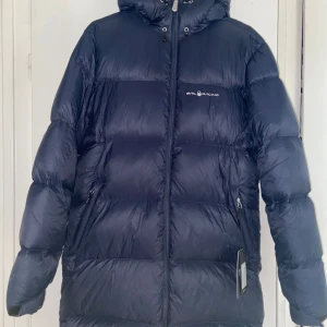 Marinblå Gravity Down Parka Sail Racing - Säljer en helt ny marinblå Gravity Down Parka från Sail Racing med prislappen. Jackan är lång, dunfylld och har huva samt dragkedja framtill. Snygg logga på bröstet och innerfoder i grått. Perfekt för kalla dagar när du vill hålla stilen och värmen.