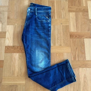 Blå Replay Anbass jeans 30/30 - Snygga blå Replay Anbass jeans med klassisk femficksdesign och diskreta slitningar. Jeansen har normal passform och raka ben, med tydliga Replay-detaljer på fickorna och bak med logopatch. Perfekta för dig som gillar stilrena och bekväma jeans.