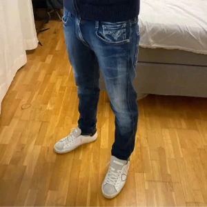 Blå Replay Anbass jeans herr - Snygga blå Replay Anbass jeans med coola slitningar och klassisk femficksdesign. Jeansen har normal passform och raka ben, med tydliga detaljer på fickorna och logga på bakfickan. Perfekta för dig som gillar en avslappnad men stilren look.
