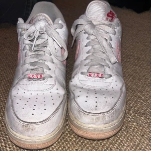Nike Air Force 1 Love Edition sneakers - Unika Nike Air Force 1 sneakers i vitt skinn med rosa detaljer och texten LOVE/NIKE på snörningen. Swoosh-loggan är rosa med mönster och skon har klassisk perforerad tå och platt sula. Perfekta för dig som vill sticka ut med en personlig touch.