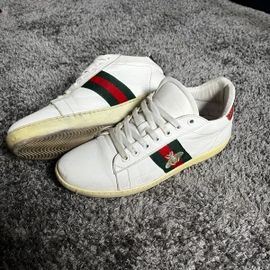 Gucci ace bee - Snygga vita sneakers från Gucci i skinn med klassiska gröna och röda ränder på sidan samt broderad guldfärgad bi. Skorna har snörning och platt sula, samt Gucci-logga under sulan. Perfekta för dig som vill sticka ut med lyxig streetstyle.