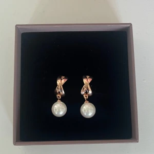 Rosefärgade örhängen med pärla - Snygga örhängen i rosefärg med tvistad design och hängande vit pärla. Perfekta för att ge en elegant touch till din look. De är lätta att bära och passar till många olika stilar.