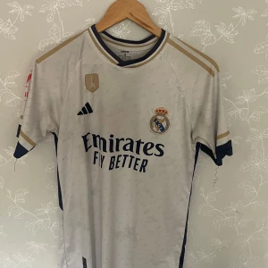 Real Madrid Rodrygo matchtröja Adidas S - Säljer en officiell Real Madrid fotbollströja från Adidas med Rodrygo och nummer 11 på ryggen. Tröjan är vit med mörkblå och guldiga detaljer, korta ärmar och tillverkad i lätt AEROREADY-material. Klubbmärke, sponsortryck och La Liga-märke på ärmen.