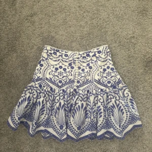 Vit och blå mönstrad kjol från Zara -  kort kjol från Zara i vitt och blått med mönster och volangkant. Kjolen har knappar framtill och en  vågig nederkant. Själv har jag aldrig använt den då den är för stor för mig tyvärr