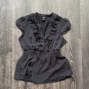 Blå prickig blus  - Supersöt svart blus från H&M med vita prickar, volangdetaljer längs v-ringningen och små knappar framtill. Blusen har korta puffärmar och smockad midja som ger en snygg siluett. Perfekt för dig som gillar retrovibbar och lekfull stil.