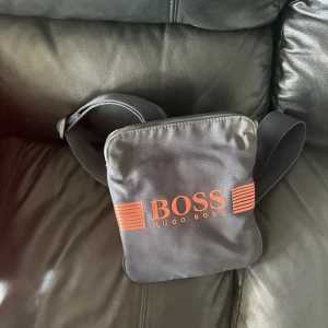 Svart axelväska från Hugo Boss - Snygg svart axelväska från Hugo Boss med röd logga och ränder framtill. Väskan har justerbar axelrem och är tillverkad i slitstark polyester. Perfekt storlek för mobil, plånbok och andra småsaker. Enkel och stilren design som passar till det mesta.