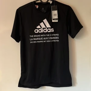 Svart Adidas t-shirt med tryck - Svart t-shirt från Adidas med stort vitt logotryck och text framtill samt tre klassiska ränder på ryggen. Tillverkad i återvunnen polyester med AEROREADY-teknologi som andas. Perfekt för sport eller chill. Klassisk passform och korta ärmar.