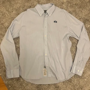 Ljusblå skjorta från La Martina M - Snygg ljusblå skjorta från La Martina med klassisk krage och broderad logga med två polospelare på bröstet. Skjortan har långa ärmar, knappar framtill och är tillverkad i mjuk bomull. Perfekt för dig som gillar stilren och sportig look.