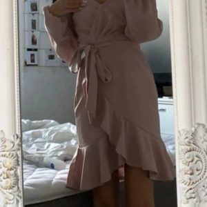 Ljusrosa omlottklänning NLY Trend - Supersöt ljusrosa omlottklänning från NLY Trend med volangkant och knyt i midjan. Klänningen har långa ärmar och v-ringning, perfekt för dig som gillar romantisk och feminin stil.