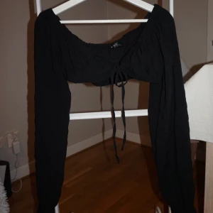 Svart croppad bolerotopp med knyt - Svart bolerotopp med lång ärm och croppad passform. Toppen har en snygg offshoulder-skärning och knytning framtill som ger en cool och trendig vibe. Perfekt att styla med höga jeans eller kjol för en edgy look.