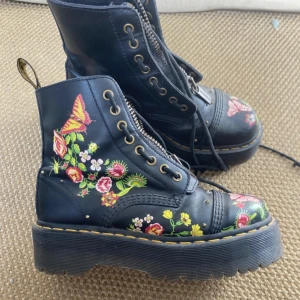 Äkta Dr. Martens Sinclair Bloom boots 39 - Svarta Dr. Martens Sinclair Bloom boots med färgglada blommor och fjärilar. Grov platt sula, snörning och dragkedja framtill. Klassisk gul söm runt sulan och robust skinnmaterial. Säljer då det inte är min stil. Använda typ en gång så de är bara lite slitna på sulan.  