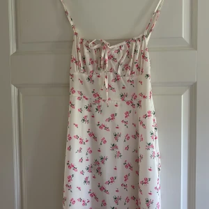 Vit blommig långklänning med snörning - Supersöt långklänning i vit med små rosa blommor och gröna blad. Klänningen har smala axelband, snörning framtill och en lätt, flowy passform. Perfekt för varma dagar och ger en romantisk vibe. Materialet känns svalt och mjukt mot huden.