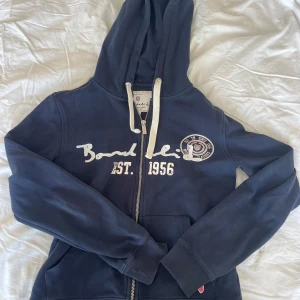 Mörkblå hoodie från Bondelid XS - Mörkblå hoodie från Bondelid med vit dragkedja och snörning i huvan. Tryck och broderad logga på bröstet, samt fickor framtill. Skön bomullskvalitet och klassisk passform, perfekt för chill dagar.